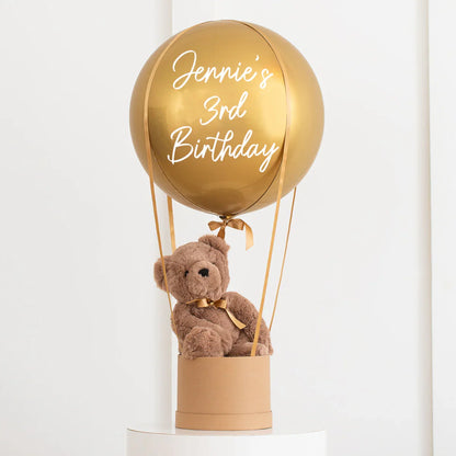Gold hot air balloon balloon with "Jennie's 3rd Birthday" text and a teddy bear inside a round gift box