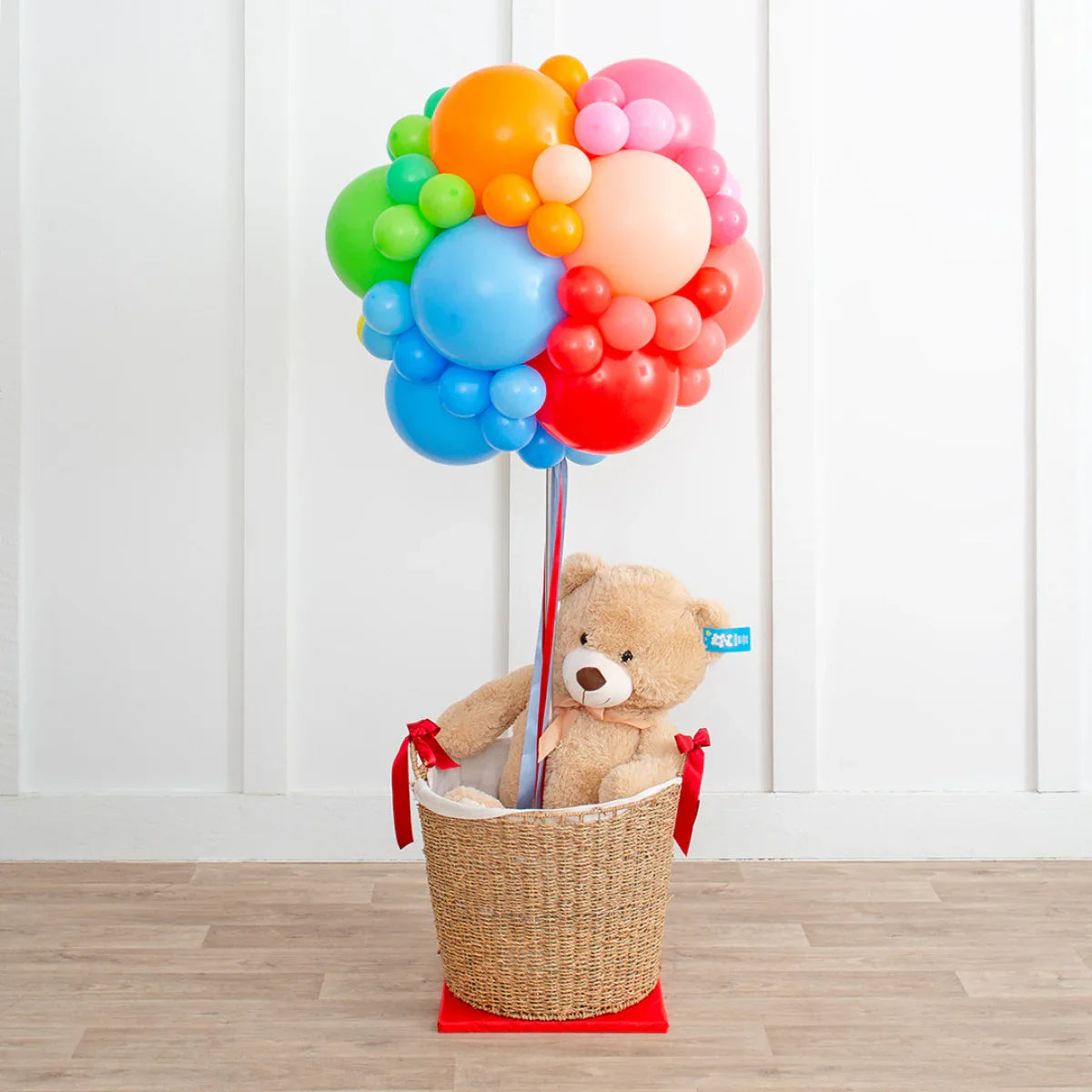 Teddy bear in wicker basket with rainbow colorful balloon cluster in white room