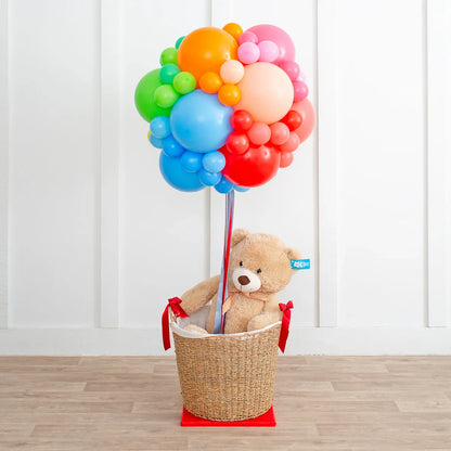 Teddy bear in wicker basket with rainbow colorful balloon cluster in white room