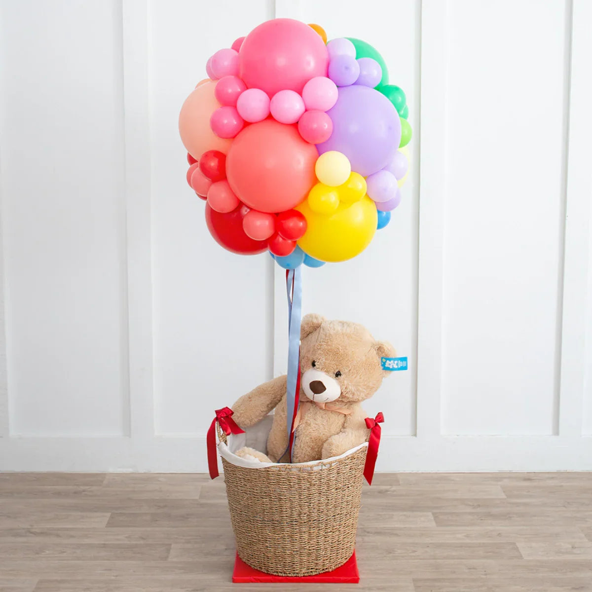 Colorful balloon bouquet attached to a wicker basket holding a teddy bear on wooden floor