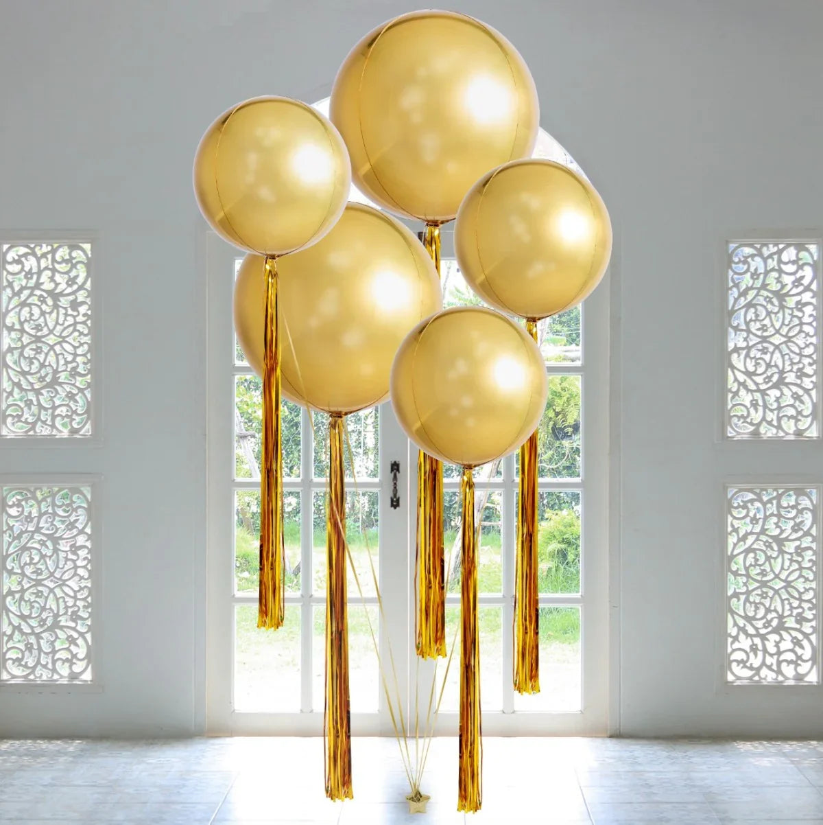 Cluster of five large gold helium balloons with gold tassels in bright white room with decorative window panels