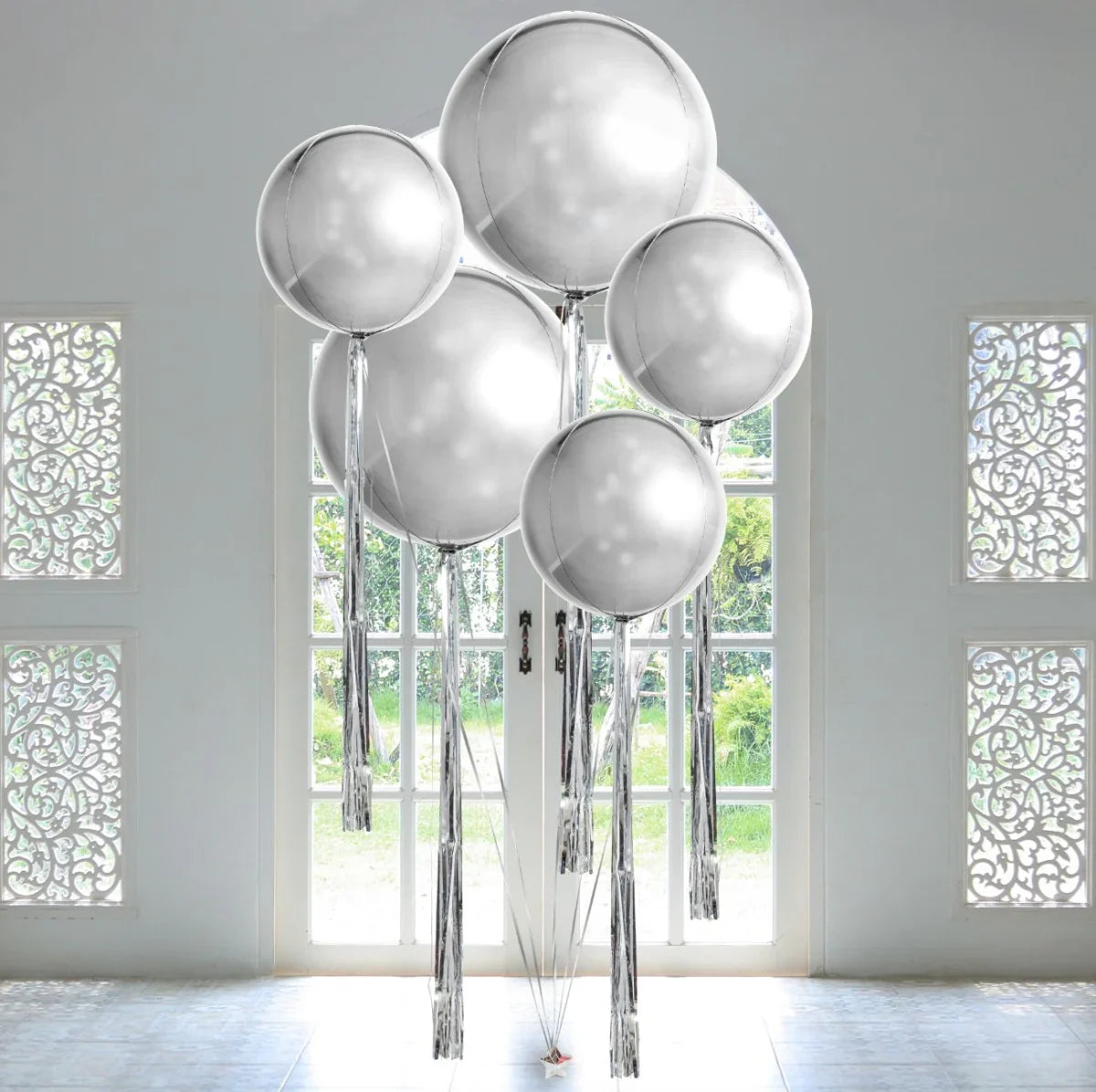 Cluster of large silver foil balloons with tassels in bright room with decorative windows